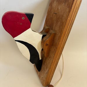 May include: A vintage wooden woodpecker toy with a red head, black beak, and white and black body. The bird is attached to a wooden board with a string and a wooden bead.
