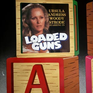 May include: Wooden toy blocks with a movie poster featuring Ursula Andress, Woody Strode, and the title "Loaded Guns." The letter "A" is prominently displayed on a block below the poster, with the name "Andress" beneath it.