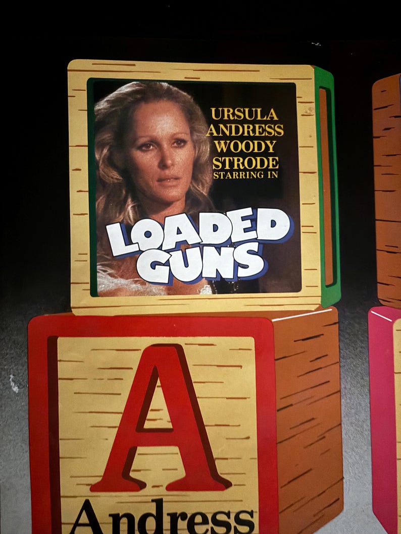 May include: Wooden toy blocks with a movie poster featuring Ursula Andress, Woody Strode, and the title "Loaded Guns." The letter "A" is prominently displayed on a block below the poster, with the name "Andress" beneath it.