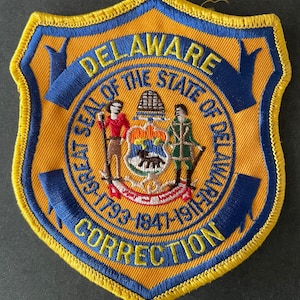 May include: A yellow and blue embroidered patch with the words "DELAWARE CORRECTION" and the seal of the state of Delaware. The seal features a farmer, a miller, and a ship.