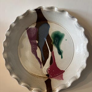 May include: A ceramic plate with a white, scalloped rim and abstract, colorful designs. The plate features a brown vertical stripe and splashes of purple, blue, green, and red. The plate has a diameter of approximately 10 inches.
