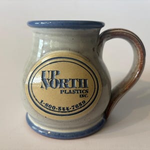 May include: A ceramic mug with a light gray body, blue rim, and blue base. It features a central oval medallion in tan with the blue text UP NORTH PLASTICS INC. and the phone number 1-800-544-7659. The mug has a curved handle with brown tones.
