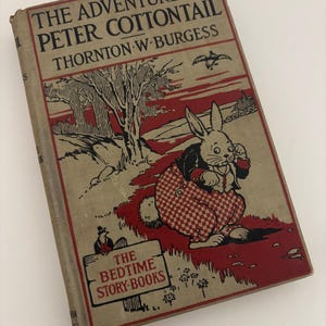 May include: Vintage hardcover book titled "The Adventures of Peter Cottontail" by Thornton W. Burgess. The cover features a red and tan illustration of a rabbit in a checkered outfit. The book is part of "The Bedtime Story-Books" series.