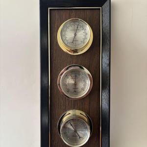 May include: A rectangular wooden weather station with three circular gauges. The top gauge is a thermometer, the middle is a barometer, and the bottom is a hygrometer. The gauges have gold and silver metal rims and are set against a dark wood background. A black frame surrounds the gauges.