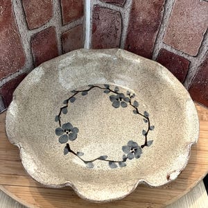 May include: A round ceramic bowl with a speckled beige finish and a scalloped edge. The interior features a hand-painted dark gray floral wreath design consisting of small blossoms and thin stems arranged in a circular pattern on the base.