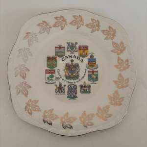 May include: A white decorative plate with a scalloped edge and gold trim. The plate features gold maple leaf designs around the rim and a central design with the word "CANADA" and various provincial coats of arms.