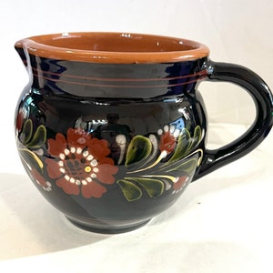 May include: A black ceramic pitcher with a brown rim and a handle. The pitcher is decorated with red, white, and green floral designs.