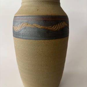 May include: A cylindrical stoneware vase with a speckled tan finish. The upper section features a wide, dark charcoal band decorated with a horizontal, carved wave pattern that reveals the lighter clay color beneath the dark surface glaze.