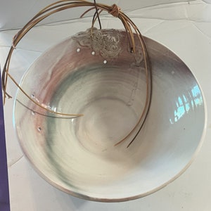 May include: A hanging ceramic planter with a glossy white interior and a color gradient of pink, green, and purple on the exterior. The planter has three drainage holes and is suspended by brown wire.