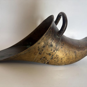 May include: Antique brass scoop with a handle. The scoop has a wide, curved shape with a long, flat base and a rounded handle. The brass has a warm, golden hue with darker areas, suggesting age and use. The scoop's design is functional and decorative.
