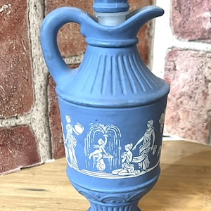 May include: A blue ceramic pitcher with a handle and a decorative stopper. The pitcher features white relief artwork depicting classical figures and a fountain scene. The design is reminiscent of antique pottery.