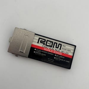 May include: A Casio ROM Pack RO-551, a vintage sound media cartridge. The black and silver cartridge features the text "WORLD SONGS" and song titles in multiple languages. The top has a silver metal cover. "NOT FOR SALE" is printed on the bottom.