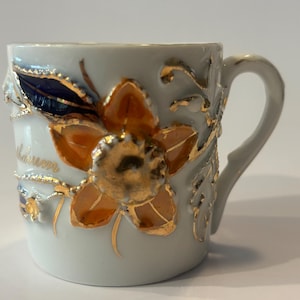 May include: A white ceramic mug featuring a raised, three-dimensional orange flower with gold-painted accents and a dark blue leaf. The design includes delicate gold-painted vines and stems wrapping around the exterior surface of the mug.