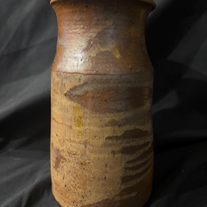 May include: A cylindrical ceramic vase with a flared rim, featuring a mottled brown and tan glaze with visible dark speckles and horizontal brushstroke textures across the surface, creating a varied earthy appearance throughout the exterior.