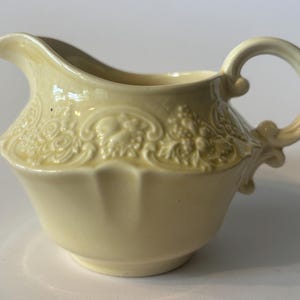 May include: A pale yellow ceramic creamer featuring a raised relief pattern of fruit and foliage around the upper body. The vessel has a curved handle, a wide pouring spout, and a smooth, glossy finish that reflects light across its surface.