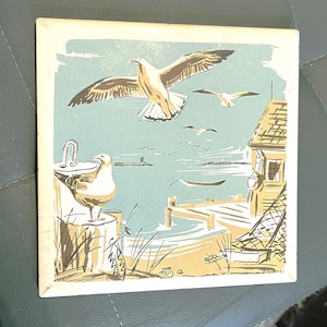 May include: Square tile featuring a coastal scene with seagulls, a pier, and a building. The artwork uses a limited color palette of beige, blue, and white. One seagull is perched on a post, while others fly in the sky. The tile has a vintage aesthetic.
