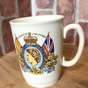 May include: Cream-colored ceramic mug featuring a commemorative design. The mug displays a portrait of a woman, a crown, flags, and the text "CORONATION JUNE 2ND 1953" and "ELIZABETH II". The mug has a curved handle and a slightly flared shape.
