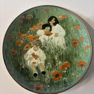 May include: A round decorative plate featuring an illustration by Jessie Willcox Smith of three children in white clothing sitting and standing in a field of green grass and orange poppy flowers, finished with a thin gold and blue rim border.
