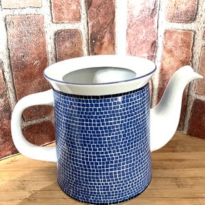 May include: A white and blue ceramic teapot with a mosaic tile pattern. The teapot has a curved spout, a handle, and a lid. The body of the teapot is covered in small blue squares, creating a textured appearance.