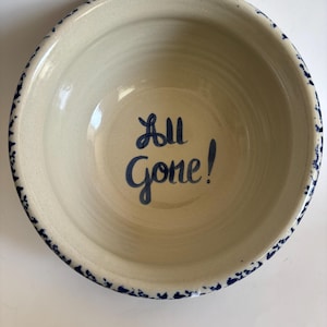 May include: Off-white ceramic bowl with a blue speckled rim and the words "All Gone!" in blue script. The bowl has a simple, rustic design, suitable for serving food or as a decorative piece.
