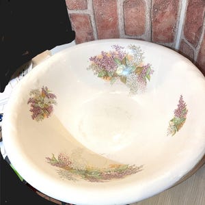 May include: Off-white, oval-shaped ceramic basin with floral lilac designs in shades of purple, white, and green. The basin has a wide rim and a smooth, glossy finish, perfect for decorative or practical use.