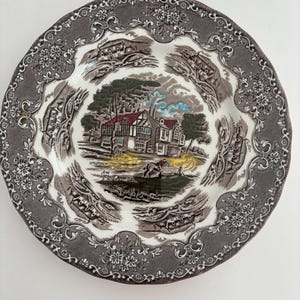 May include: A round, decorative plate with a detailed landscape scene in the center. The plate features a brown and white color scheme with a floral border. The central image depicts a building with a red roof, trees, and a person on a horse.