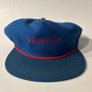 May include: A blue baseball cap featuring the word Monticello embroidered in red thread on the front panel. The cap includes a red braided cord trim across the base of the crown where it meets the flat brim, and a matching red button on top.