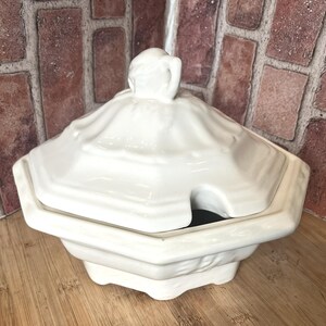 May include: A white ceramic soup tureen with a lid and a handle. The tureen has a geometric shape with a scalloped edge and a small opening for a ladle. The background includes a brick wall and a wooden surface.