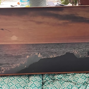May include: A rectangular wall clock with a coastal sunset scene. The clock face features a seascape with a dark rocky coastline, seagulls, and a setting sun. The clock hands are silver, and the numbers are red.