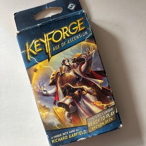 May include: A KeyForge Age of Ascension card game box. The box is blue with gold and white text and a fantasy illustration of a character in armor. The box reads "A Unique Deck Game by Richard Garfield" and "Contains 1 Unique Ready-to-Play Archon Deck."