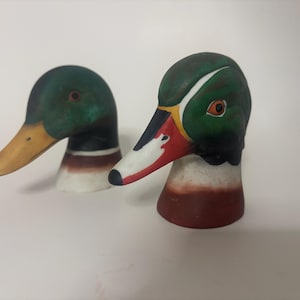 May include: Two decorative duck head sculptures. The ducks have green heads, yellow beaks, and red and white accents. The sculptures are painted with detailed features and sit on a white surface.