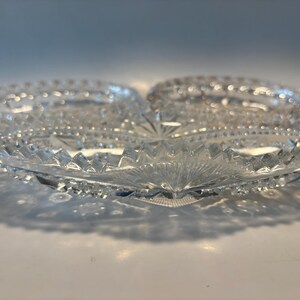 May include: A clear, three-sectioned glass serving dish. The dish has a starburst design in the center and a decorative, textured rim. The glass is cut with geometric patterns, creating a sparkling effect. The dish casts a shadow on the white surface.