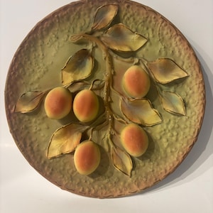 May include: A round decorative wall plate featuring a raised, three-dimensional relief of a branch with four orange-toned fruits and several light green leaves. The background of the plate has a textured, mottled olive green and brown finish.