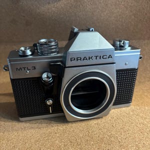 May include: A silver and black Praktica MTL3 camera. The camera has a silver body with black textured grips and a large lens opening. The word "PRAKTICA" is printed in black on the front of the camera. The camera is on a brown surface.