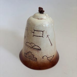 May include: A ceramic bell with a cream-colored body and a brown base. The bell features etched designs of geometric shapes and a bird. A small, dark brown piece is attached to the top.