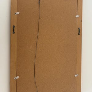 May include: Back of a rectangular picture frame with a light brown wooden frame. The backing is a corkboard material. The frame has metal hardware and a wire for hanging. White plastic clips secure the backing.