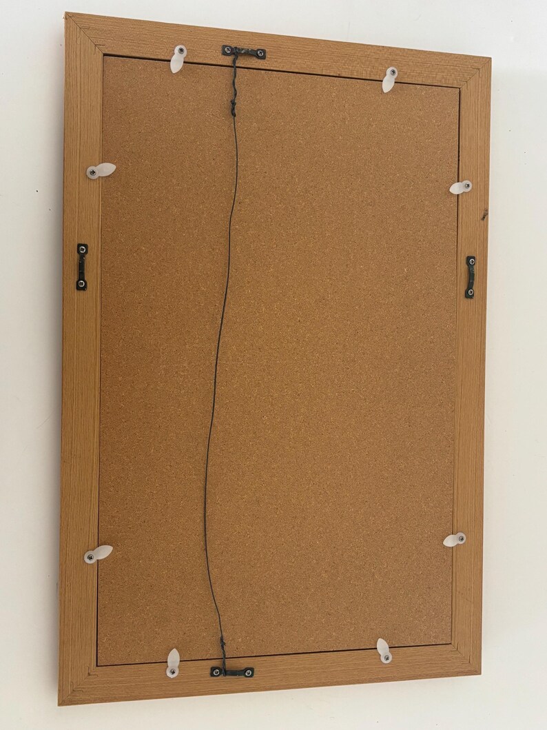 May include: Back of a rectangular picture frame with a light brown wooden frame. The backing is a corkboard material. The frame has metal hardware and a wire for hanging. White plastic clips secure the backing.