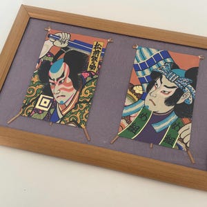 May include: Framed artwork featuring two Japanese Kabuki actor prints. The prints are colorful, with detailed illustrations of the actors' faces and costumes. The frame is made of light brown wood, and the background is a muted purple.