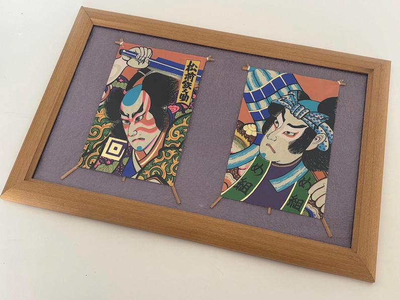 May include: Framed artwork featuring two Japanese Kabuki actor prints. The prints are colorful, with detailed illustrations of the actors' faces and costumes. The frame is made of light brown wood, and the background is a muted purple.