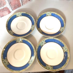 May include: Four white ceramic saucers with a blue border and floral accents. The saucers have a gold trim and a central indentation for a teacup. The design is symmetrical and elegant, suitable for a tea set.