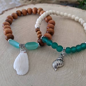 May include: Two bracelets with wooden and white beads, accented with green glass beads and shell charms. One charm is a white seashell, the other is a silver seashell.