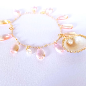 Könnte beinhalten: Ein zartes goldfarbenes Armband mit tropfenförmigen rosa- und pfirsichfarbenen Anhängern. Ein Muschelanhänger mit einer Perle ist ebenfalls angebracht. Das Armband befindet sich auf weißem Hintergrund.