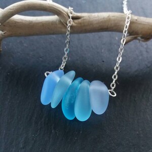 Op de afbeelding: Een zilveren ketting met zes blauwe zeeglas hangers in verschillende tinten blauw. De druppelvormige hangers zijn in een rij gerangschikt. De ketting wordt getoond tegen een donkere achtergrond.