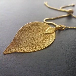 Könnte beinhalten: Eine goldfarbene Blatt-Anhänger-Halskette. Der Anhänger hat die Form eines detaillierten Blattes mit komplizierten Adern, und ein kleinerer Blatt-Anhänger ist befestigt. Die Halskette hat eine zarte Kette mit einer verstellbaren Gleitperle.