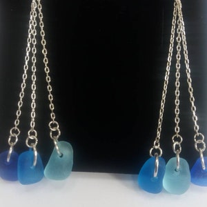 May include: A pair of dangle earrings with three silver chains and blue sea glass charms. The sea glass is in shades of blue, teal, and aqua.
