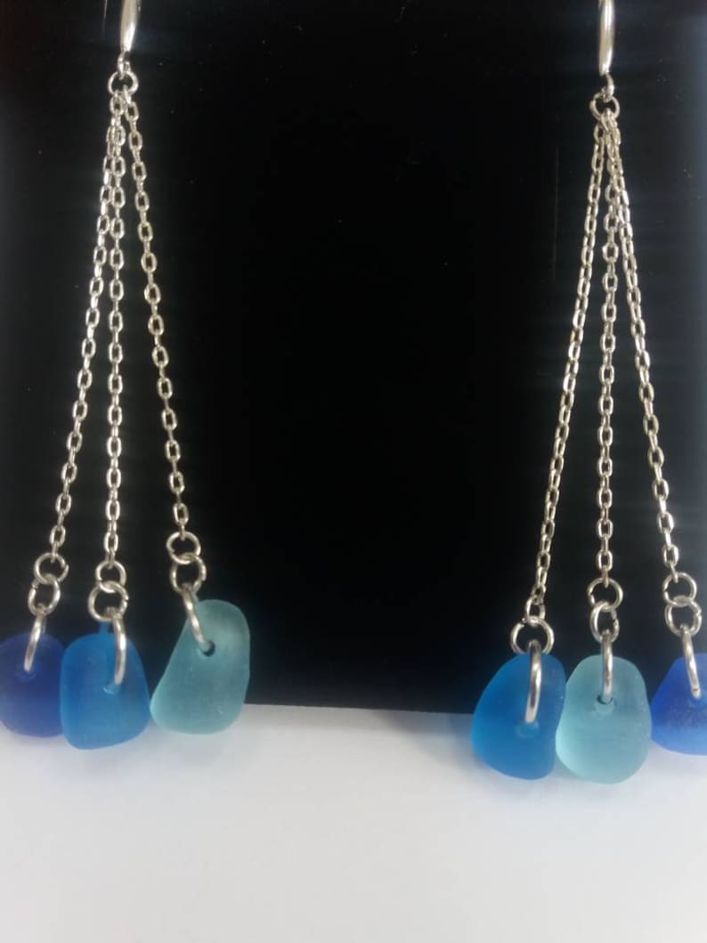 May include: A pair of dangle earrings with three silver chains and blue sea glass charms. The sea glass is in shades of blue, teal, and aqua.