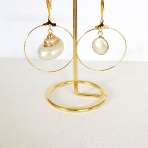 May include: A pair of gold hoop earrings with a white shell pendant on each hoop. The earrings are displayed on a gold stand.