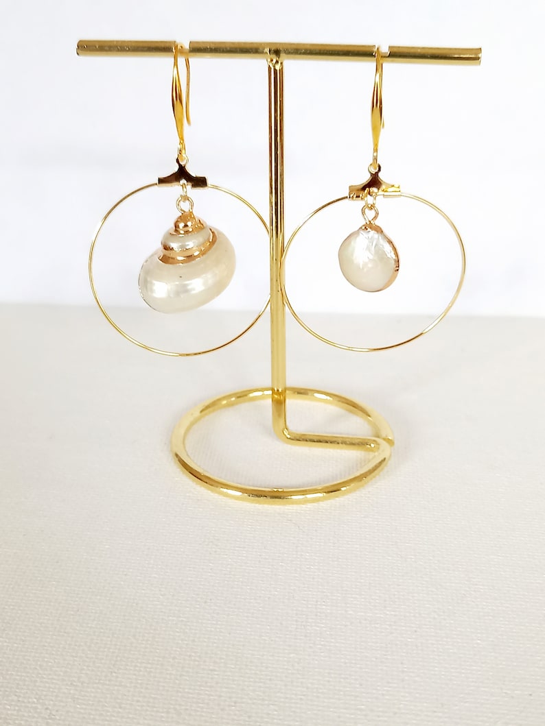May include: A pair of gold hoop earrings with a white shell pendant on each hoop. The earrings are displayed on a gold stand.