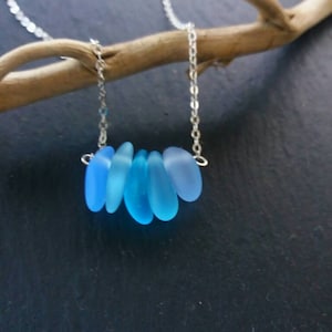 Op de afbeelding: Een delicate ketting met een fijne zilveren ketting, met een reeks afgestudeerde, lichtblauwe zeeglasstukken. Het zeeglas is geregen. De ketting wordt gepresenteerd tegen een donkere achtergrond, die de koele tinten van het zeeglas benadrukt.