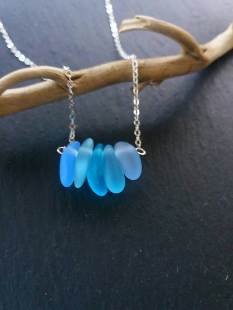Op de afbeelding: Een delicate ketting met een fijne zilveren ketting, met een reeks afgestudeerde, lichtblauwe zeeglasstukken. Het zeeglas is geregen. De ketting wordt gepresenteerd tegen een donkere achtergrond, die de koele tinten van het zeeglas benadrukt.